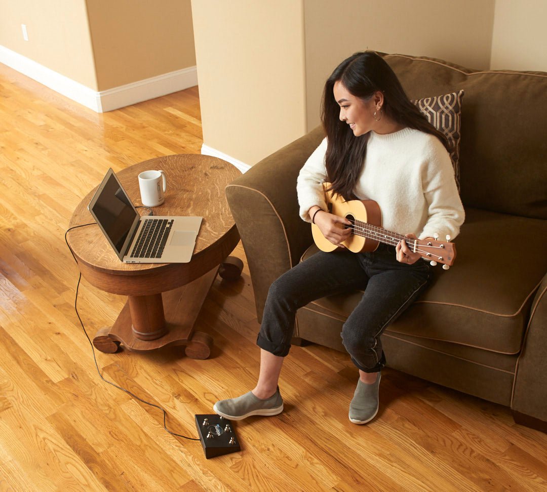 A young girl is learning Ukulele online  by looping and slowing down sections of the video without taking her hands off her instrument, saving time and without being frustrated.  Her foot is on the Vidami YouTube looper which is  connected to a laptop computer via USB cable.